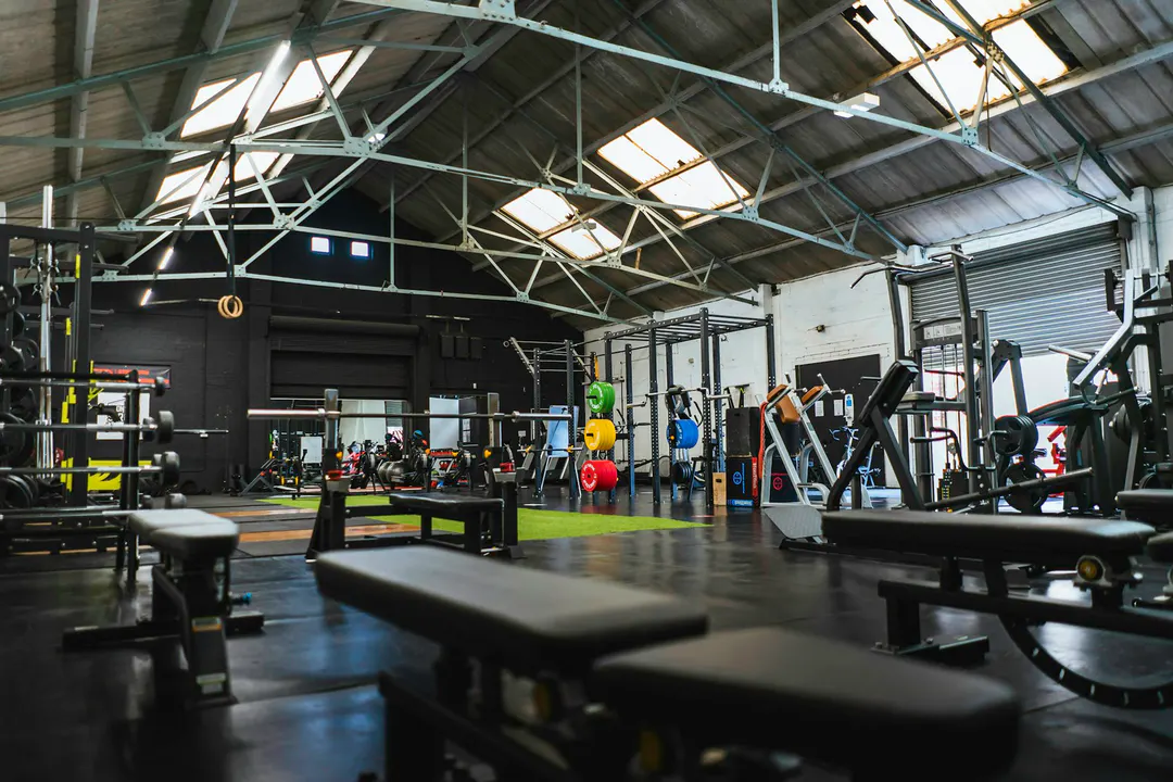 Intérieur d'une salle d'entraînement fonctionnel aménagée dans un hangar industriel, avec bancs, racks à squat, haltères et grande verrière au plafond