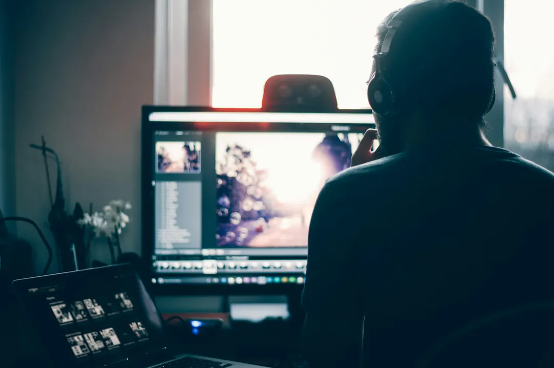 Photographe de dos, casque sur les oreilles, retouchant une série de photos sur un écran d'ordinateur près d'une fenêtre lumineuse, un ordinateur portable affichant des vignettes au premier plan
