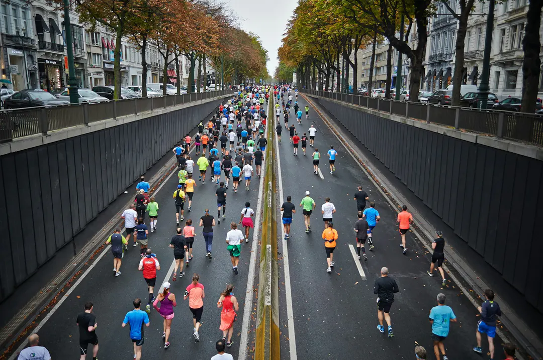 Marathon en ville vu d'en haut : des centaines de coureurs de dos descendent une trémie routière bordée d'arbres aux couleurs d'automne (Photo : Mārtiņš Zemlickis / Unsplash)
