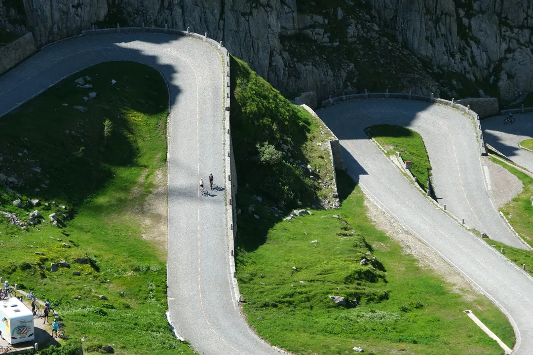 Route de col en lacets sur un versant herbeux, avec des cyclistes en pleine ascension au pied d'une falaise