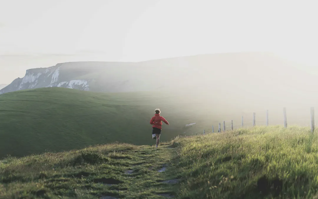 Traileur seul courant sur un sentier de crête herbeux dans une lumière dorée, falaises et brume en arrière-plan