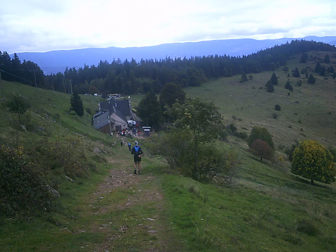 Sentier herbeux qui descend vers une ferme d'alpage où est installé le ravitaillement d'un trail, quelques coureurs de dos sur le chemin et des montagnes boisées à l'horizon