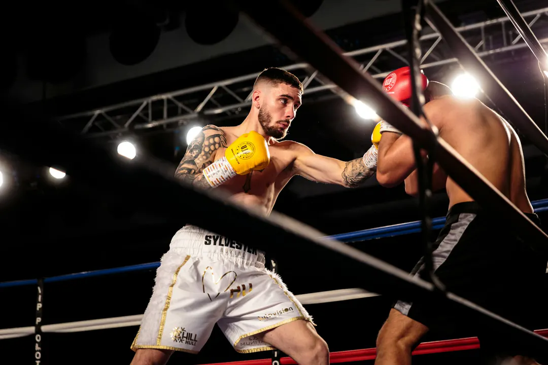 Boxeur en short blanc et gants jaunes lançant un direct, photographié à travers les cordes du ring sous les projecteurs d'un gala