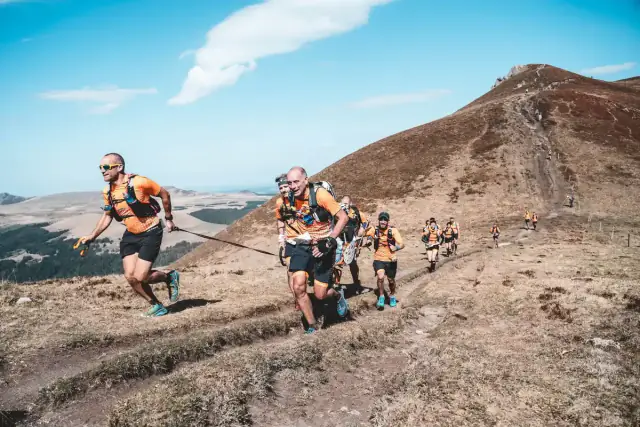 Coureurs de trail en maillot orange dans une montée herbeuse, sous un ciel bleu